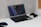 A sleek laptop is open on a white desk, displaying a minimalist wallpaper with geometric shapes and vibrant lines. To the right of the laptop, a black gaming controller rests, while to the left, a glowing computer mouse is visible. The desk setup suggests a blend of work and play in a modern setting, with a corkboard partially visible on the wall.