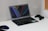 A sleek laptop is open on a white desk, displaying a minimalist wallpaper with geometric shapes and vibrant lines. To the right of the laptop, a black gaming controller rests, while to the left, a glowing computer mouse is visible. The desk setup suggests a blend of work and play in a modern setting, with a corkboard partially visible on the wall.