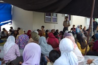 A group of people are gathered indoors, seated on the floor, some wearing headscarves. The setting appears informal, with people engaged in conversation or listening. There is a man standing, possibly addressing the group, and a mix of men, women, and children present.