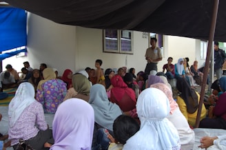 A group of people are gathered indoors, seated on the floor, some wearing headscarves. The setting appears informal, with people engaged in conversation or listening. There is a man standing, possibly addressing the group, and a mix of men, women, and children present.