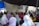 A group of people are gathered indoors, seated on the floor, some wearing headscarves. The setting appears informal, with people engaged in conversation or listening. There is a man standing, possibly addressing the group, and a mix of men, women, and children present.