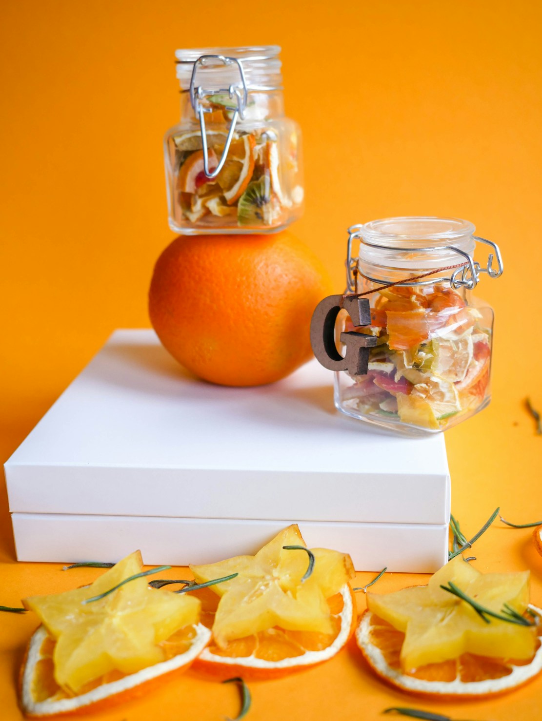 A composition featuring dried fruit slices and two glass jars filled with dried fruit, set against an orange background. An orange fruit supports the top jar, while star fruit slices and orange slices are arranged on a white box and the surface below.