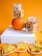 A composition featuring dried fruit slices and two glass jars filled with dried fruit, set against an orange background. An orange fruit supports the top jar, while star fruit slices and orange slices are arranged on a white box and the surface below.