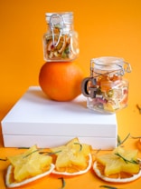 A composition featuring dried fruit slices and two glass jars filled with dried fruit, set against an orange background. An orange fruit supports the top jar, while star fruit slices and orange slices are arranged on a white box and the surface below.
