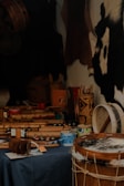 A variety of musical instruments with traditional designs are placed on a dark tablecloth. The focus is on drums and shakers made from wood and hide, with intricate carvings and patterns. Some paint cans and other tools are also visible, suggesting a craftsman's workshop. The background features animal hides hanging on the wall.