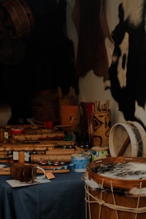A variety of musical instruments with traditional designs are placed on a dark tablecloth. The focus is on drums and shakers made from wood and hide, with intricate carvings and patterns. Some paint cans and other tools are also visible, suggesting a craftsman's workshop. The background features animal hides hanging on the wall.