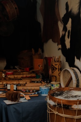 A variety of musical instruments with traditional designs are placed on a dark tablecloth. The focus is on drums and shakers made from wood and hide, with intricate carvings and patterns. Some paint cans and other tools are also visible, suggesting a craftsman's workshop. The background features animal hides hanging on the wall.