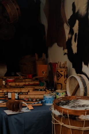 A variety of musical instruments with traditional designs are placed on a dark tablecloth. The focus is on drums and shakers made from wood and hide, with intricate carvings and patterns. Some paint cans and other tools are also visible, suggesting a craftsman's workshop. The background features animal hides hanging on the wall.