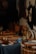 A variety of musical instruments with traditional designs are placed on a dark tablecloth. The focus is on drums and shakers made from wood and hide, with intricate carvings and patterns. Some paint cans and other tools are also visible, suggesting a craftsman's workshop. The background features animal hides hanging on the wall.