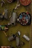 A collection of metal objects hung on a wire mesh display, including tin cans with designs, cheese graters, and decorative clocks. The arrangement presents a variety of upcycled or recycled items with rustic and industrial aesthetics.