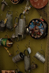 A collection of metal objects hung on a wire mesh display, including tin cans with designs, cheese graters, and decorative clocks. The arrangement presents a variety of upcycled or recycled items with rustic and industrial aesthetics.