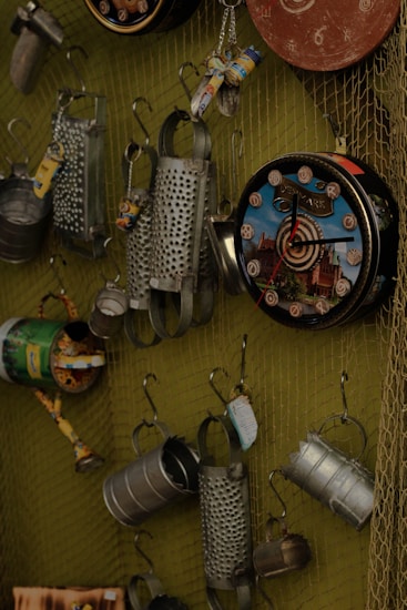 A collection of metal objects hung on a wire mesh display, including tin cans with designs, cheese graters, and decorative clocks. The arrangement presents a variety of upcycled or recycled items with rustic and industrial aesthetics.
