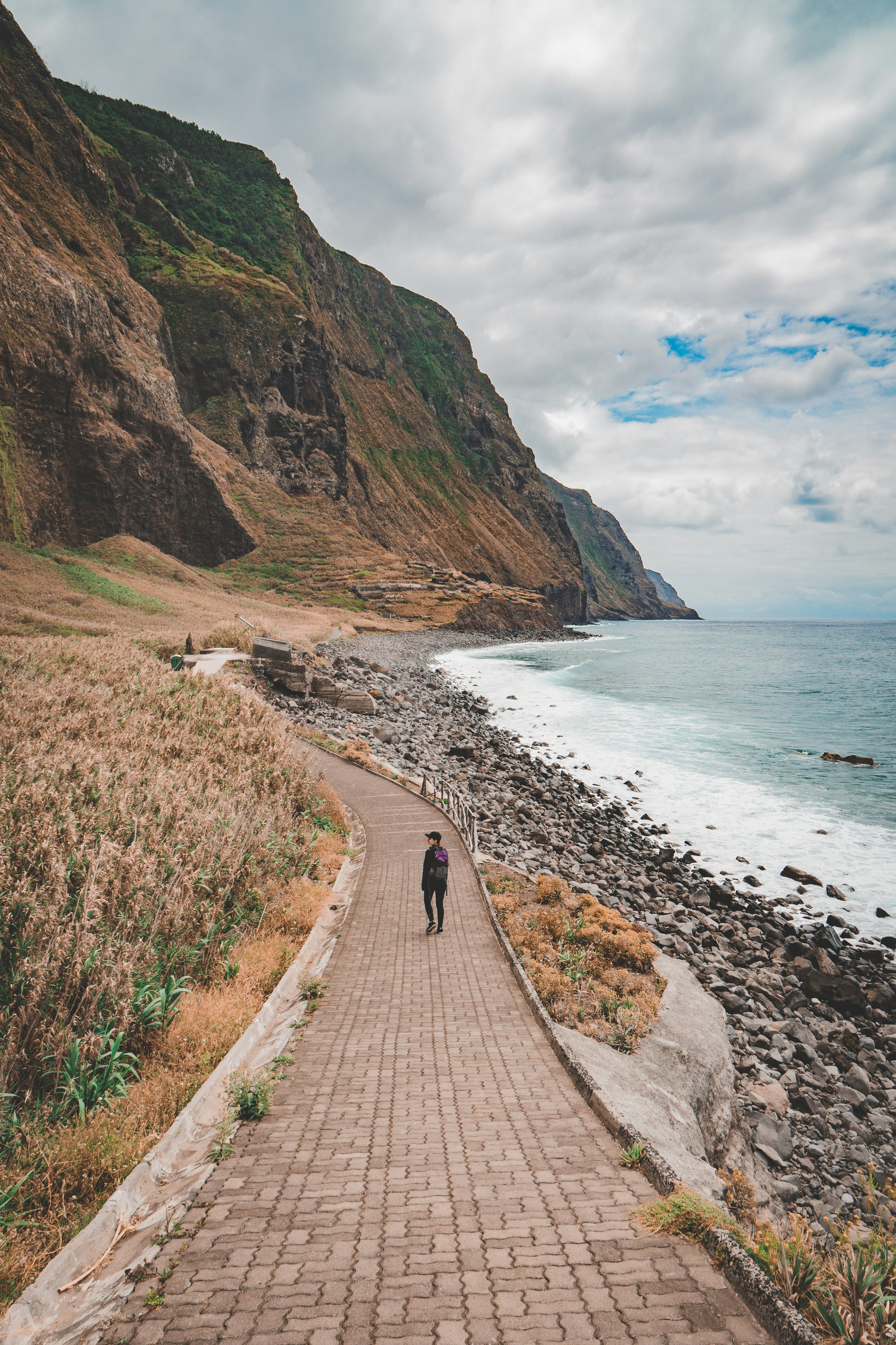 person walking on road viewing mountain and sea under white and blue skies during daytime