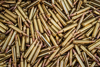 A close-up view of a large pile of brass-colored rifle cartridges with copper-colored bullet tips, arranged in a random, overlapping pattern.