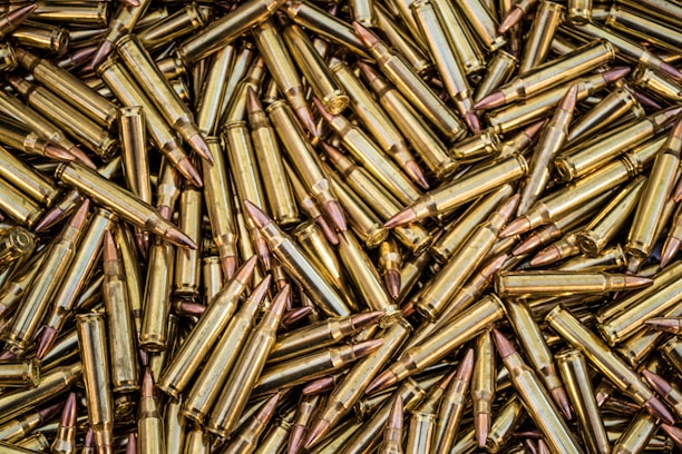 A close-up view of a large pile of brass-colored rifle cartridges with copper-colored bullet tips, arranged in a random, overlapping pattern.