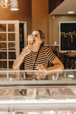 A person is enjoying a large ice cream cone inside a warmly lit ice cream shop. They are standing behind a display case filled with various flavors of ice cream. The person is wearing a striped shirt and appears to be happy and relaxed. The shop has a cozy ambiance with warm lighting and some decorative elements visible in the background.