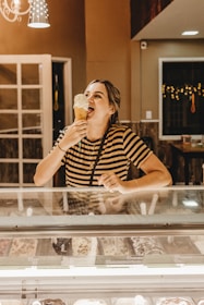 A person is enjoying a large ice cream cone inside a warmly lit ice cream shop. They are standing behind a display case filled with various flavors of ice cream. The person is wearing a striped shirt and appears to be happy and relaxed. The shop has a cozy ambiance with warm lighting and some decorative elements visible in the background.