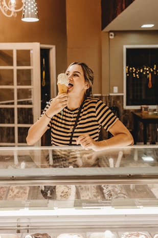 A person is enjoying a large ice cream cone inside a warmly lit ice cream shop. They are standing behind a display case filled with various flavors of ice cream. The person is wearing a striped shirt and appears to be happy and relaxed. The shop has a cozy ambiance with warm lighting and some decorative elements visible in the background.