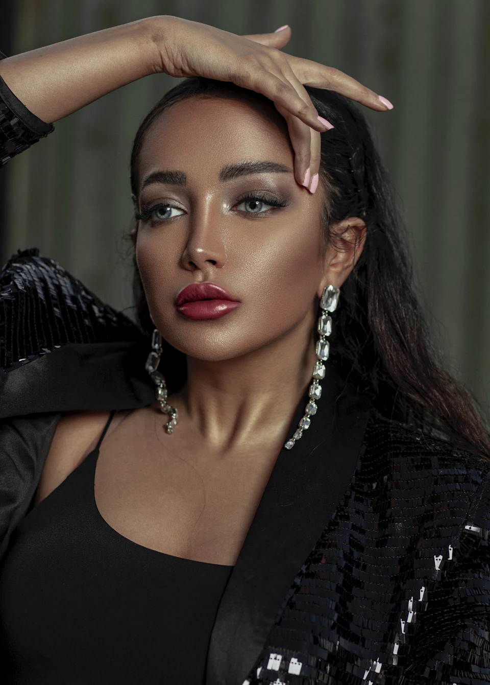 The image shows a woman with long dark hair wearing a black sequin jacket and large, dangling earrings. Her hand is raised to her face, and she has one finger resting on her forehead. The background appears to be a greenish-brown color, which contrasts with the woman's dark hair and outfit.