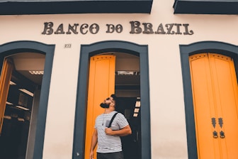 A man with a beard is standing in front of a building with the sign 'Banco do Brazil.' He is wearing a striped t-shirt and carrying a backpack over his shoulder. The building has striking yellow doors with blue-green trim.