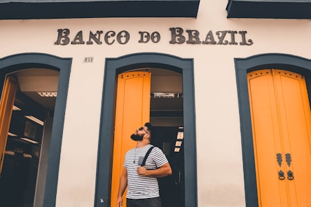 A man with a beard is standing in front of a building with the sign 'Banco do Brazil.' He is wearing a striped t-shirt and carrying a backpack over his shoulder. The building has striking yellow doors with blue-green trim.