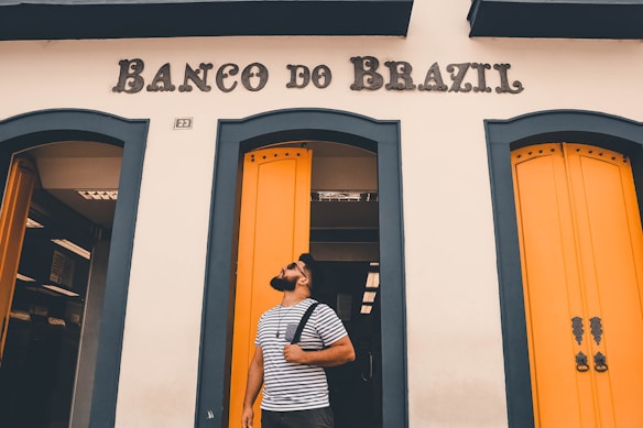 A man with a beard is standing in front of a building with the sign 'Banco do Brazil.' He is wearing a striped t-shirt and carrying a backpack over his shoulder. The building has striking yellow doors with blue-green trim.