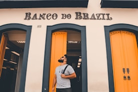 A man with a beard is standing in front of a building with the sign 'Banco do Brazil.' He is wearing a striped t-shirt and carrying a backpack over his shoulder. The building has striking yellow doors with blue-green trim.