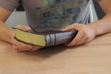 A person is holding a thick book with a brown cover embossed with the words 'The Word of God' in gold lettering. The book is being held open at the edge of a light wooden table. The person is wearing a casual gray t-shirt with a multi-colored geometric design.