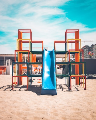 A vibrant playground structure stands on sandy ground under a clear blue sky. The structure consists of colorful metal frames in red, yellow, orange, and green, with a bright blue slide at the center. Behind the playground, there are some buildings and construction cranes, indicating an urban setting. Outdoor furniture and orange parasols are visible in the background.