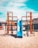 A vibrant playground structure stands on sandy ground under a clear blue sky. The structure consists of colorful metal frames in red, yellow, orange, and green, with a bright blue slide at the center. Behind the playground, there are some buildings and construction cranes, indicating an urban setting. Outdoor furniture and orange parasols are visible in the background.