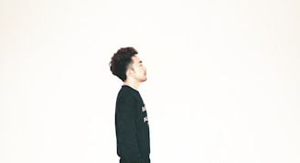 A person with dark hair is wearing a black shirt and facing sideways against a plain, light background. The composition emphasizes simplicity and minimalism.