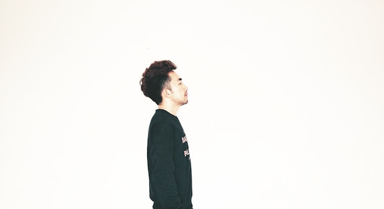 A person with dark hair is wearing a black shirt and facing sideways against a plain, light background. The composition emphasizes simplicity and minimalism.