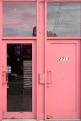 A pair of pink, rectangular doors with glass panels on either side. One door has a handle with a 'PUSH' sign, and above, a window reflects the cloudy sky and power lines. The adjacent door displays the number '2711'.
