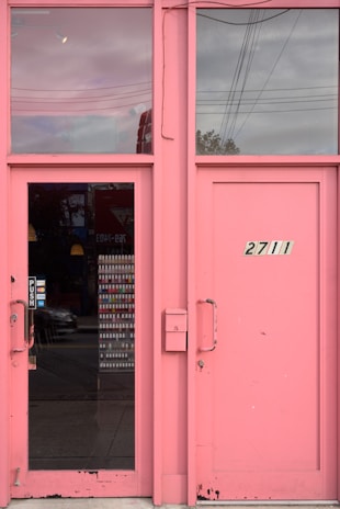 A pair of pink, rectangular doors with glass panels on either side. One door has a handle with a 'PUSH' sign, and above, a window reflects the cloudy sky and power lines. The adjacent door displays the number '2711'.