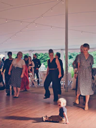 A toddler sits on a wooden floor under a large tent, surrounded by adults who are walking or standing. The lighting is warm with string lights hanging from the tent ceiling. There are white folding chairs and tables in the background, suggesting a formal or social event.
