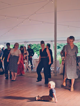A toddler sits on a wooden floor under a large tent, surrounded by adults who are walking or standing. The lighting is warm with string lights hanging from the tent ceiling. There are white folding chairs and tables in the background, suggesting a formal or social event.