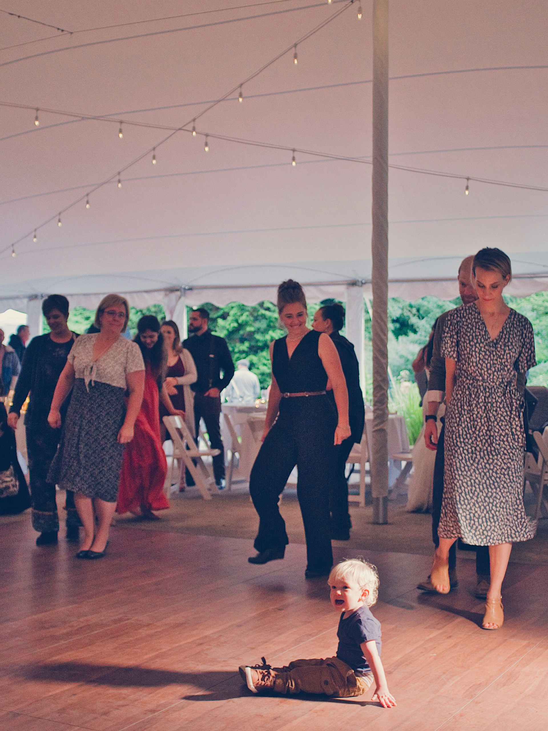 A toddler sits on a wooden floor under a large tent, surrounded by adults who are walking or standing. The lighting is warm with string lights hanging from the tent ceiling. There are white folding chairs and tables in the background, suggesting a formal or social event.