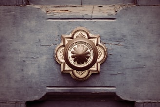 A vintage-style metal door handle is mounted on a distressed wooden door. The door handle is ornate and symmetrical, with a floral-inspired design in the center, surrounded by a decorative, circular pattern. The wood appears weathered, with visible cracks and peeling paint.
