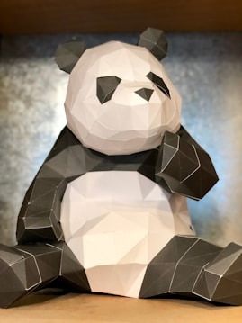 A paper-crafted geometric panda bear sculpture is sitting upright. The panda is composed of black and white polygonal shapes with distinct angular lines, giving it a modern and artistic appearance. The background is a neutral, slightly textured surface.