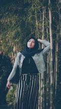 A person wearing a hijab and denim jacket stands among tall bamboo plants, looking upward with a thoughtful expression. They are dressed in striped pants and carrying a black bag, with one hand resting on their head.