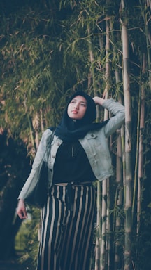 A person wearing a hijab and denim jacket stands among tall bamboo plants, looking upward with a thoughtful expression. They are dressed in striped pants and carrying a black bag, with one hand resting on their head.