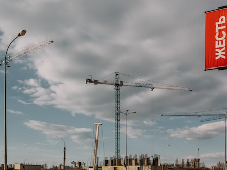 Several construction cranes are positioned against a backdrop of scattered clouds and a mostly blue sky. A red sign with Cyrillic text hangs prominently on the right side of the image. The scene is an urban construction site with partially constructed buildings visible in the lower part of the image.