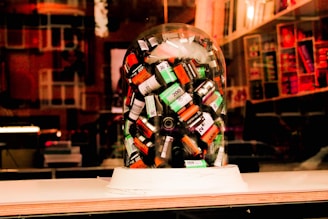 A collection of various film canisters is enclosed under a transparent glass dome, displayed in what appears to be a store window. The background features shelves filled with various colorful items, and the scene is imbued with warm lighting, suggesting an inviting atmosphere often found in photography or art stores.