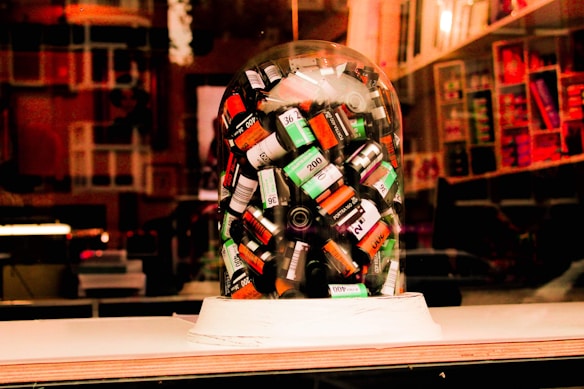 A collection of various film canisters is enclosed under a transparent glass dome, displayed in what appears to be a store window. The background features shelves filled with various colorful items, and the scene is imbued with warm lighting, suggesting an inviting atmosphere often found in photography or art stores.