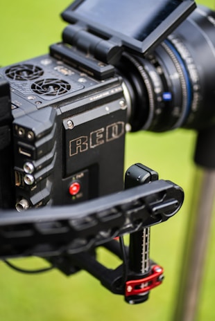 Close-up view of a professional RED camera with visible buttons, HDMI port, lens, and display screen on a green blurred background.
