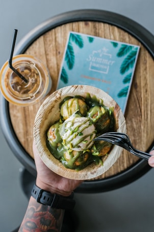 A hand holding a bowl filled with dessert that includes green sauce and a dollop of cream, garnished with pistachio pieces. In the background, a wooden round table holds an iced drink with a black straw and a teal-colored menu with palm leaf illustrations.