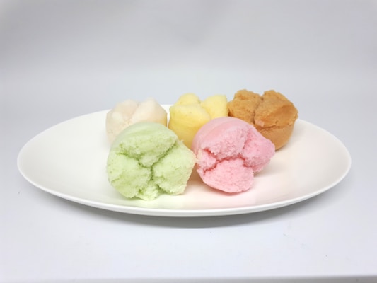 Five colorful steamed muffins are arranged on a white plate. The muffins feature light green, pink, yellow, beige, and brown colors, each appearing fluffy and soft.