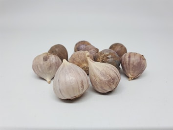 A cluster of garlic bulbs displayed on a clean, light-colored surface. The garlic bulbs have a natural, papery outer skin with a mix of off-white and purple hues.