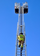 A firefighter is climbing a tall metal ladder on a fire truck, reaching towards two platforms against a clear blue sky. The firefighter is wearing protective gear, including a helmet and high-visibility jacket.