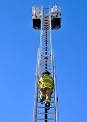 A firefighter is climbing a tall metal ladder on a fire truck, reaching towards two platforms against a clear blue sky. The firefighter is wearing protective gear, including a helmet and high-visibility jacket.
